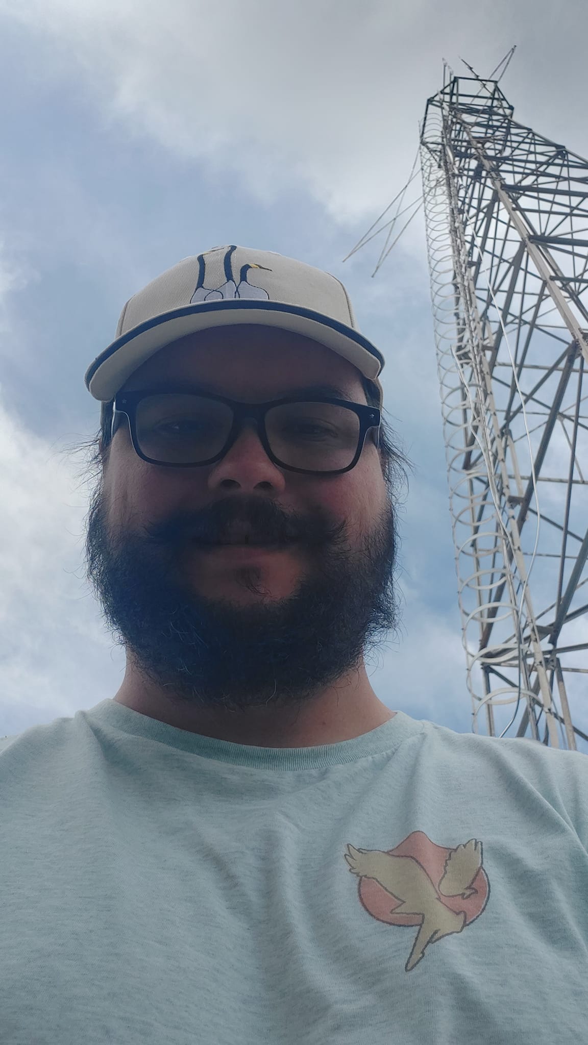 A man short hair and bushy facial hair stands in front of an old radio tower